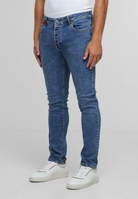 Egy férfi karcsú kék farmernadrágot és fehér sneakers-t visel, egy sima világosszürke háttér előtt áll.