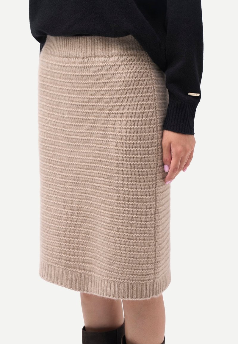 Beige stickad knälång kjol kombinerad med svart tröja och stövlar, sedd från sidan med ribbad midjerem och fåll.