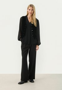 Outfit nero composto da una blusa trasparente a maniche lunghe sovrapposta a un gilet di velluto. I pantaloni neri dal taglio morbido e gli stivaletti neri alla caviglia completano il look.