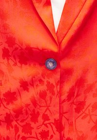 Φωτεινό πορτοκαλί σακάκι με διακριτικά λουλουδάτα σχέδια, ένα διακοσμητικό ασημένιο κουμπί, φορεμένο πάνω από ένα λευκό ριμπ μπλουζάκι.