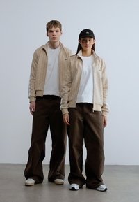 Μπεζ ζακέτες με μοτίβα λοξόν κλείσιμο πάνω από λευκά πουκάμισα, συνδυασμένα με φαρδιά καφέ παντελόνια. Μαύρο καπέλο φορεμένο από έναν, και οι δύο με ουδέτερα παπούτσια. Μινιμαλιστικό φόντο.