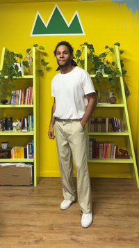 Lahke bež hlače z gladkim teksturo, bela majica s kratkimi rokavi in beli supergi. Zeleno poudarjen zid z režami za knjige in rastlinami v ozadju.