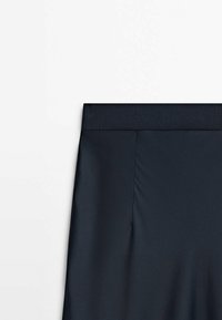 Jupe noire avec une large ceinture côtelée et un tissu lisse, montrée de la taille vers le bas sur un fond blanc uni.