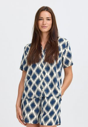 Žena s dlhými hnedými vlasmi, oblečená v modro-bielom vzorovanom krátkoročkovom košeľovom tričku a zodpovedajúcich šortkách, stojaca s jednou rukou v vrecku.