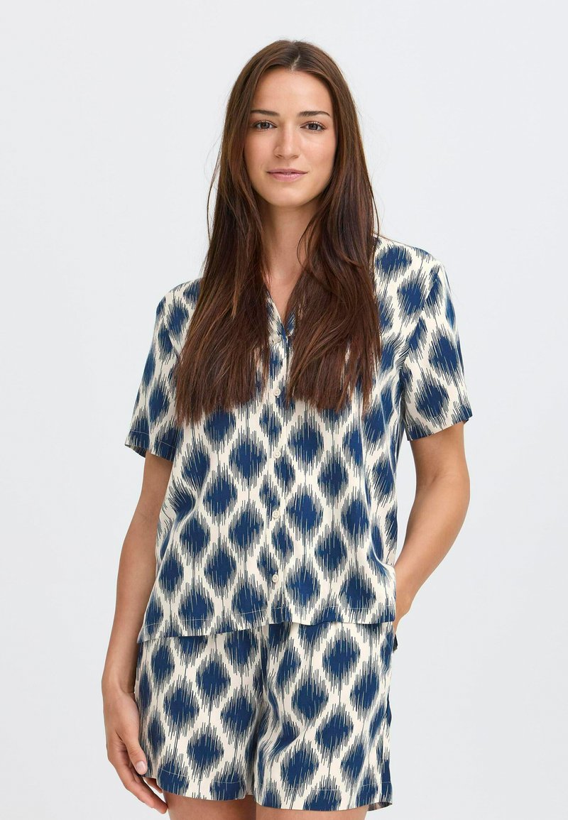 Žena s dlhými hnedými vlasmi, oblečená v modro-bielom vzorovanom krátkoročkovom košeľovom tričku a zodpovedajúcich šortkách, stojaca s jednou rukou v vrecku.