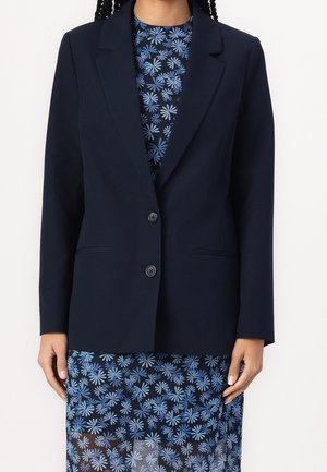 Personne portant un blazer bleu marine sur une robe bleu foncé avec un motif floral bleu clair, debout devant un fond blanc uni.