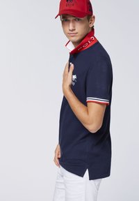 Navy Poloshirt mit rot-weißer gestreifter Kragen und Bündchen, ausgestattet mit einem bestickten Logo. Getragen mit weißen Hosen und einer roten Mütze.