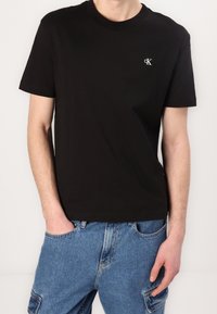 Hombre con camiseta de manga corta negra con pequeño logo blanco "cK" en el pecho y pantalones cargo azules, con las manos en los bolsillos.