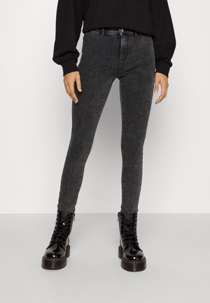 Personne portant un jean noir ajusté et délavé, un sweat-shirt noir et des bottes de combat noires brillantes à lacets, debout sur un sol gris clair.