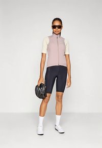 Žena u biciklističkoj odjeći s svijetloružastim prslukom, crnim šortsevima, bijelim čarapama i cipelama, drži crnu kacigu i nosi sunčane naočale.