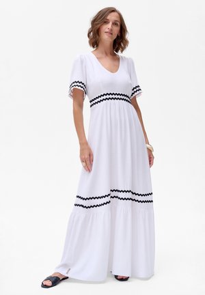 Witte maxi-jurk met V-hals, zwarte zigzagborduring op de mouwen en taille, en een gelaagde zoom. Gemaakt van lichte stof.