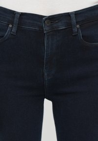 Tuvuma tumšzilas džinsu jostas daļa ar pogu, rāvējslēdzēju, jostu cilpiņām un priekšējām kabatām.