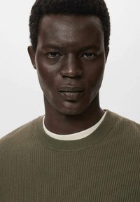 Olivgrön ribbad tröja med rundad halsringning, lagd över en vit t-shirt, med en strukturerad stickning och en klassisk passform.