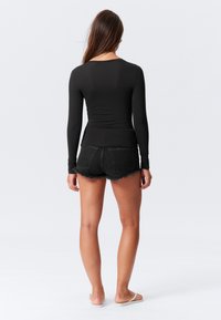 Černý top s dlouhým rukávem a přiléhavým střihem, doplněný o černé džíny s třepením. Modelka má na sobě lehké sandály, pohled zezadu.