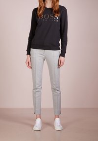 Svart sweatshirt med "BOSS"-logotyp i metallisk tryck, kombinerad med ljusgrå, smala byxor med sidosömmar och fållar, bärs med vita sneakers.