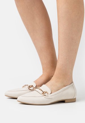 Mocassins beiges en cuir avec un talon plat, ornés d'une chaîne dorée et d'un design à bout pointu. Texture lisse.