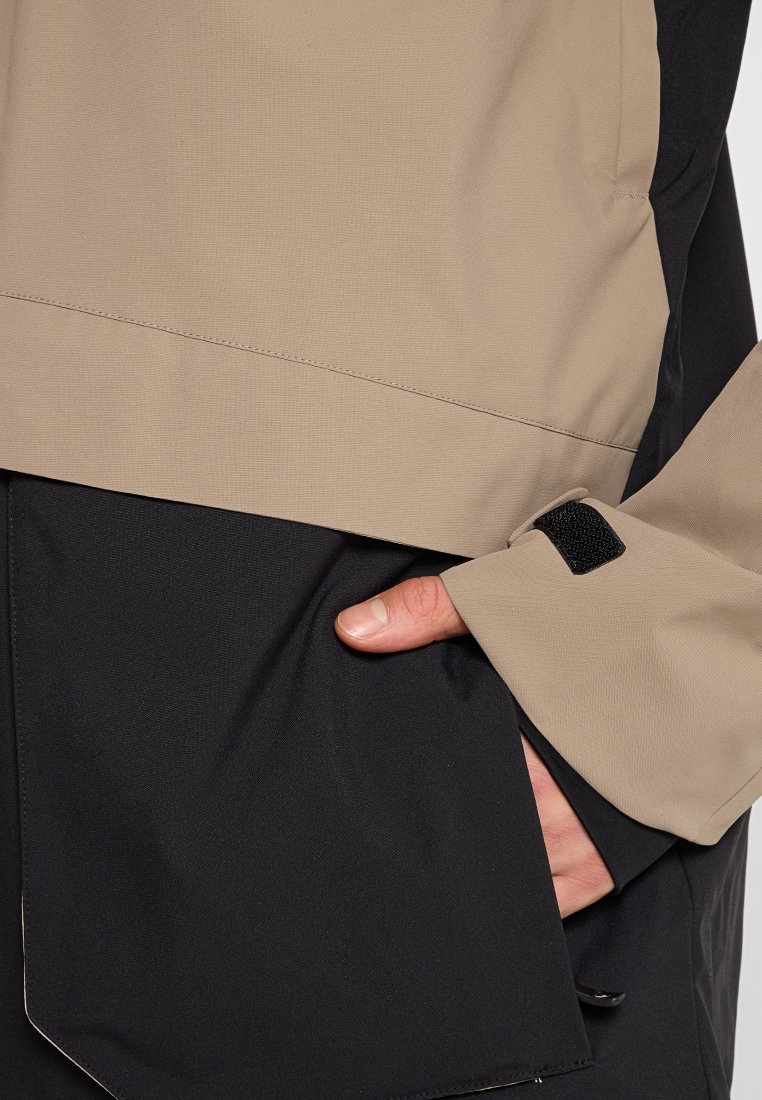 Wasserabweisende Jacke mit einem beige oberen und einem schwarzen unteren Abschnitt, strukturiertem Stoff und einem mit Klettverschluss verstellbaren Ärmelbund sowie einer Seitentasche.