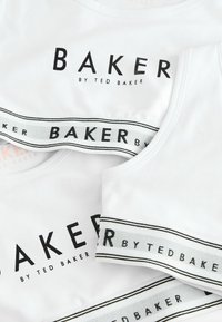 Hvide tanktops med stort sort "BAKER" logo og tekst, med en kontrasterende elastisk bånd, der viser gentaget "BAKER" i sort og grå.