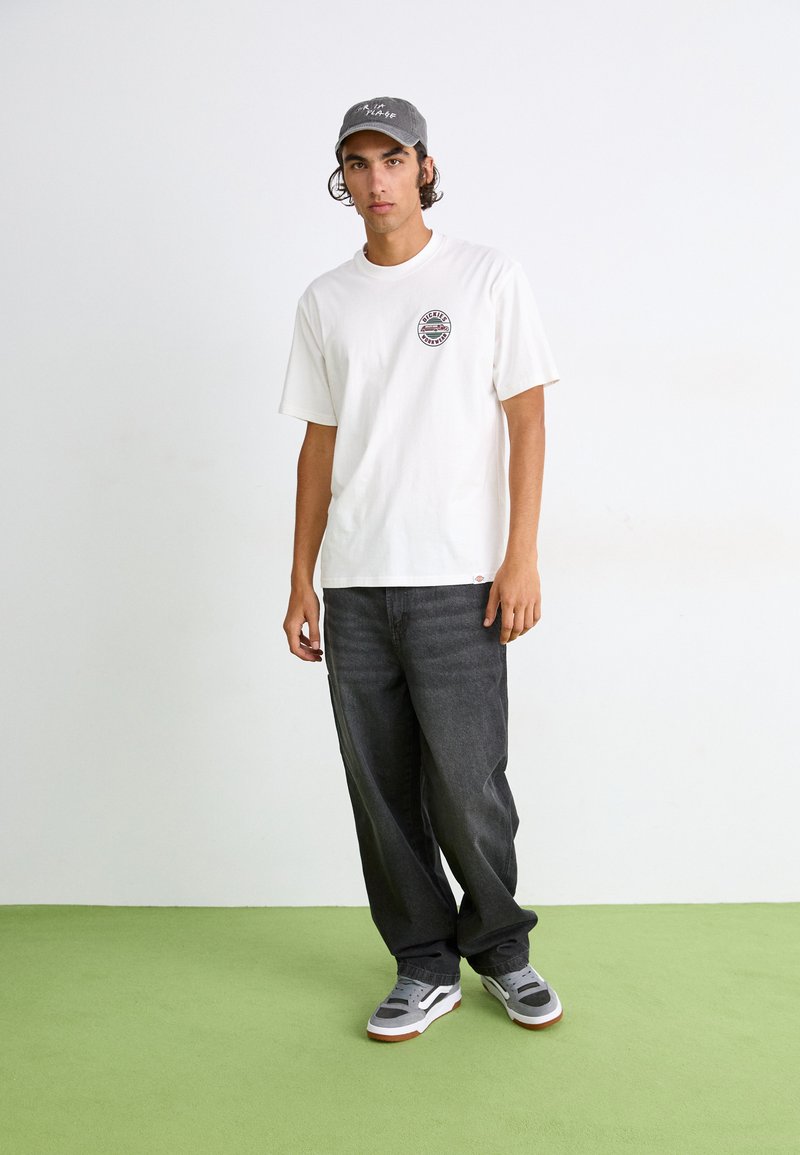 Weißes T-Shirt mit kurzen Ärmeln und rundem Logo, dunkle weite Jeans und graue Sneakers, dazu eine graue Cap. Steht auf einem grünen Teppich.