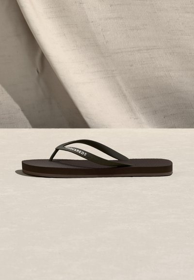 Σκούρο καφέ σανδάλι σαγιονάρας με λεπτές λουρίδες, μία από τις οποίες φέρει το κείμενο "DSQUARED2", τοποθετημένο πάνω σε φωτεινή επιφάνεια με υφασμάτινο φόντο.