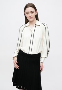 Woman with straight brown hair wearing a cream blouse with black piping and a black skirt with metal eyelet details, posing against a white background.
