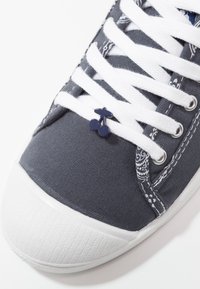 Sneaker in tela con tomaia grigia scura, stringhe bianche e punta in gomma; presenta un piccolo charm a forma di ciliegia blu sulle stringhe e dettagli decorativi.
