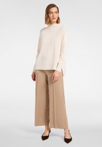 Frau trägt einen cremefarbenen Rollkragenpullover, weite beige Hose und dunkelbraune spitze Ballerinas, steht vor einem schlichten hellen Hintergrund.