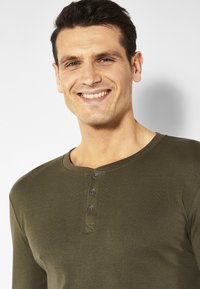 Olivgrünes Langarm-Henley-Shirt aus weichem, strukturiertem Stoff mit drei schwarzen Knöpfen am Hals und einem abgerundeten Kragendesign.