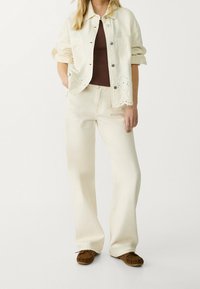 Ivoorkleurige spijkerjack met oogjesdetails aan de zoom, gecombineerd met beige broek met brede pijpen en bruine mocassinstijl schoenen.