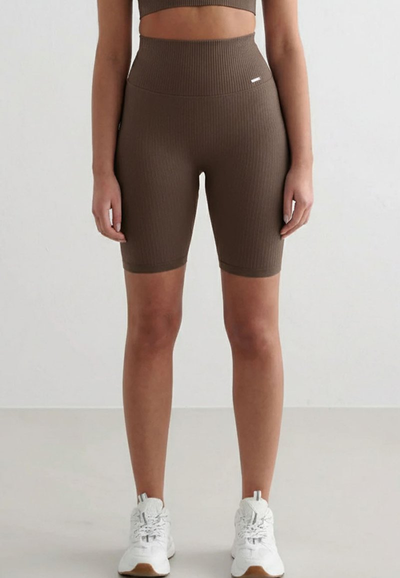 Femme portant un short cycliste marron côtelé à taille haute et des baskets blanches, debout devant un fond clair uni.