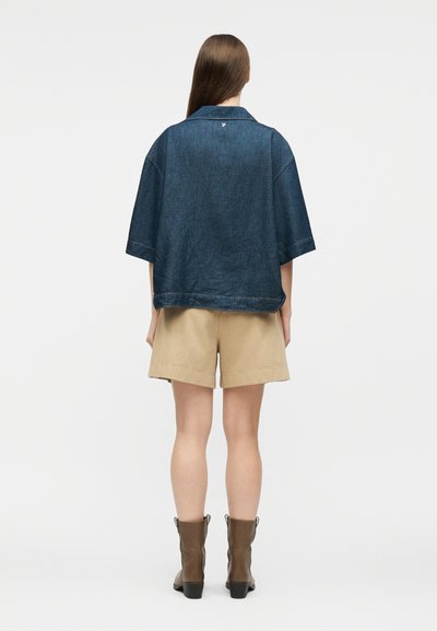 Γυναίκα με μακριά καστανά μαλλιά, φοράει φαρδιά κοντομάνικη σκουρόχρωμη denim πουκαμίσα, μπεζ σορτς και καφέ μποτάκια στον αστράγαλο, στέκεται γυρισμένη προς τα πίσω.