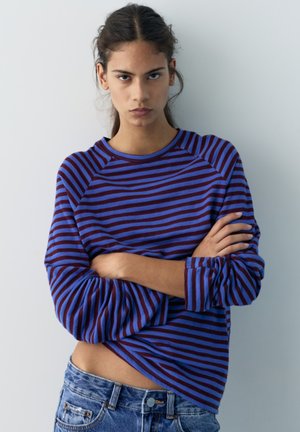 Jong persoon met gekruiste armen, gekleed in een blauw met kastanjebruin gestreept shirt met lange mouwen en een blauwe spijkerbroek, tegen een effen lichte achtergrond.