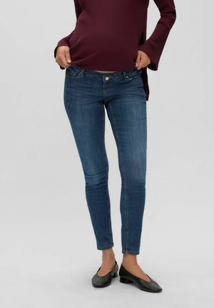 Personne portant un jean slim bleu et des chaussures noires à talons bas, tenant son haut à manches longues bordeaux près de la taille.