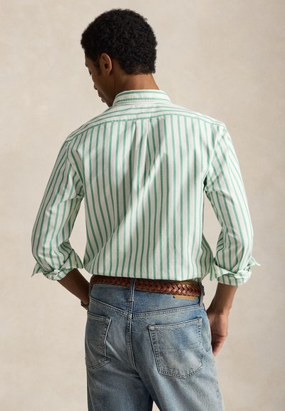 Άνδρας με κοντά σγουρά μαλλιά που φοράει λευκό πουκάμισο με πράσινες κάθετες ρίγες και ανοιχτό μπλε τζιν, γυρισμένος πλάτη με τα χέρια στη μέση.