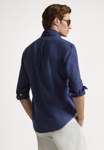 Camisa de lino azul marino con un corte relajado, mangas remangadas, detalles de botones y una pieza en la espalda, combinada con pantalones de color claro.