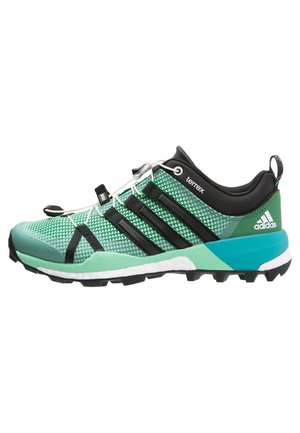 Groene en zwarte Adidas Terrex trailrunning schoen met robuuste zool, zwarte strepen en witte veters op een witte achtergrond.