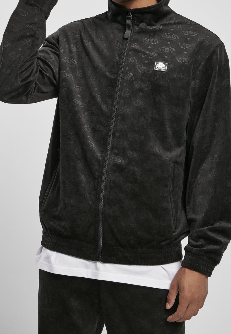 Svart zip-up jacka i mjukt texturerat tyg med ett subtilt mönster. Fitted manschetter och en zippered krage. Litet logopatch på bröstet.