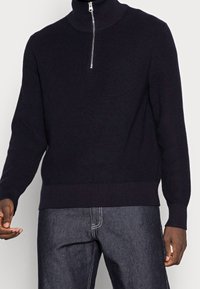 Pull côtelé bleu marine avec un col haut et une fermeture éclair argentée à mi-hauteur. Il présente des manches longues et un ourlet ajusté, associé à un jean en denim foncé.