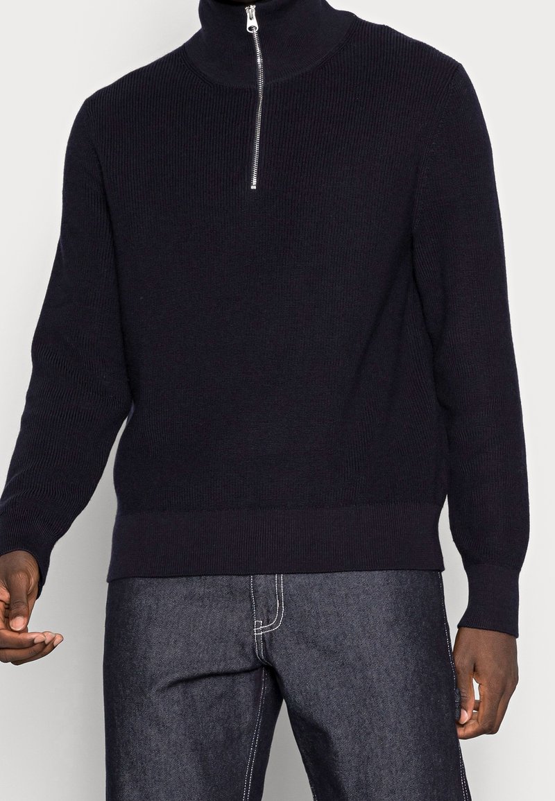 Pull côtelé bleu marine avec un col haut et une fermeture éclair argentée à mi-hauteur. Il présente des manches longues et un ourlet ajusté, associé à un jean en denim foncé.