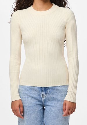 Femme portant un pull crème côtelé, ajusté et à manches longues, associé à un jean en denim bleu clair, debout devant un fond blanc uni.