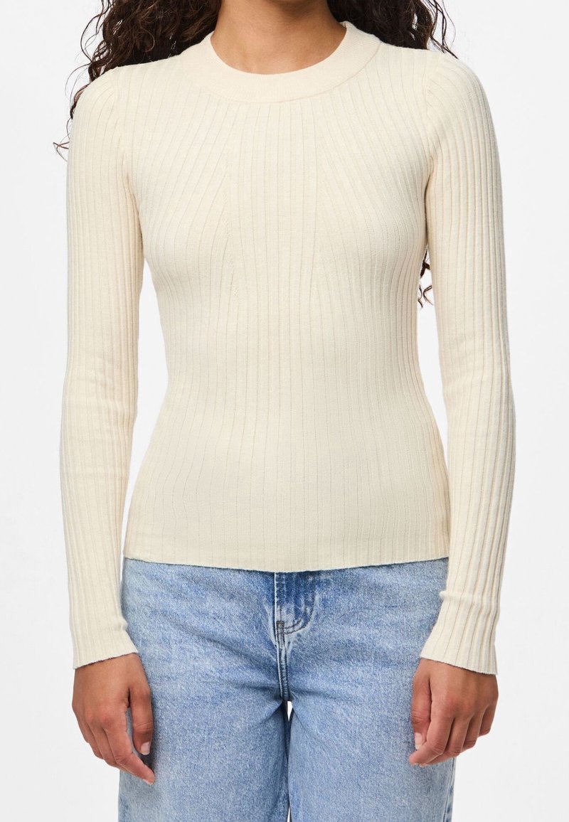 Femme portant un pull crème côtelé, ajusté et à manches longues, associé à un jean en denim bleu clair, debout devant un fond blanc uni.