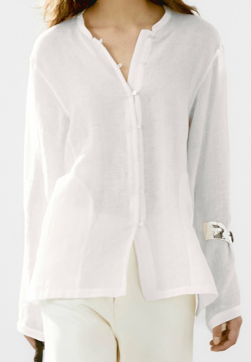Femme portant un chemisier blanc léger à manches longues boutonné avec un bracelet manchette en argent à un bras.