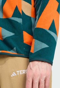 Fleece-Oberteil mit geometrischen Mustern in Blaugrün, Orange und Grau; weiche Textur; elastische Bündchen; kombiniert mit beigen Hosen mit weißem Logo.