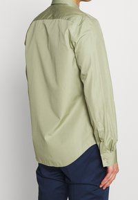 Camicia a maniche lunghe verde chiaro con colletto puntato, polsini con bottoni e orlo dritto. Tessuto liscio con poche pieghe.