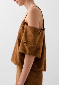Brown, perforated fabric top with a wide, off-the-shoulder design. Features floral cutouts and a smooth texture. Adjustable straps.