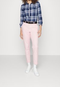 Person klädd i en blå och vit rutig skjorta instopps i ljusrosa byxor med ett svart bälte, vita strumpor och vita sneakers.