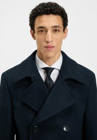 Peacoat in lana blu navy con ampi revers e chiusura doppia; indossato sopra una camicia bianca e una cravatta nera. Texture liscia.