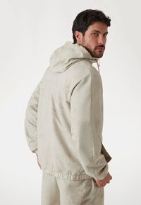 Hooded beige jacket with a subtle texture, featuring a zip closure, elastic cuffs, and a relaxed fit. Back view shows no visible logos.