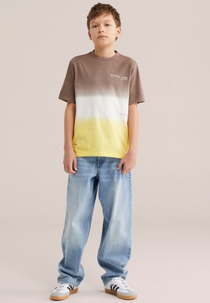 Niño de pie vistiendo una camiseta con degradado en marrón, blanco y amarillo, vaqueros holgados de color azul claro y zapatillas blancas con rayas negras.