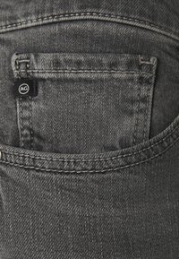 Jeans en denim gris avec une petite poche arrière portant un logo AG, des coutures visibles et une texture douce. Le tissu présente une légère usure.